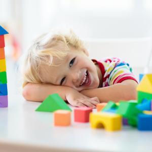 A toddler smiling at the camera while resting his head on a table filled with toy building blocks.